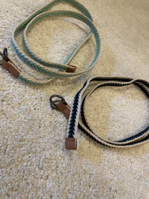 Två flätade bälten i blå och grön - Säljer två smala flätade bälten med läderdetaljer och metallspänne. Ett bälte är blått och vitt, det andra är ljusgrönt och vitt. Perfekta för att ge din outfit en snygg touch. Båda har en stilren, randig look och passar till många olika stilar.