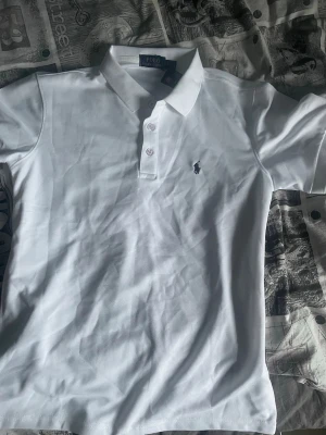 Vit pikétröja från Polo Ralph Lauren - Klassisk vit pikétröja från Polo Ralph Lauren med krage, knappar och den ikoniska broderade loggan på bröstet. Tillverkad i mjuk bomull som känns skön mot huden. Perfekt basic-plagg med snygg passform och tidlös stil.