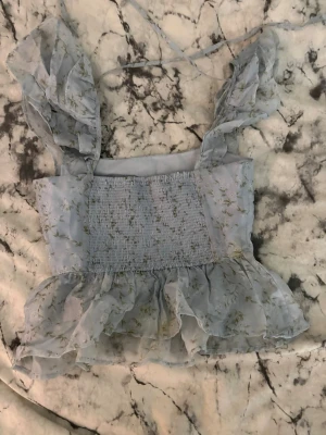 Blommig ljusblå volangtop - Söt ljusblå topp med blommigt mönster och volanger. Toppen har breda axelband med volangdetaljer och smockad framsida för en skön passform. Materialet är tunt och transparent, perfekt för sommarens varma dagar. Volang nedtill ger en romantisk vibe.