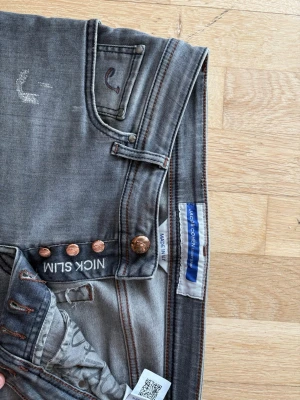 Gråa slim jeans från Jacob Cohën - Säljer ett par gråa slim fit jeans från Jacob Cohën, modell Nick cost slim. Jeansen har fem fickor, kopparfärgade knappar och diskreta slitningar för en cool look. Tillverkade i Italien och har klassiska orange sömmar cosignade med märket.