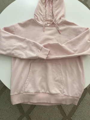 Ljusrosa hoodie med ficka - En stilren ljusrosa hoodie med huva och dragsnören. Hoodien har en stor magficka och ribbade muddar vid ärmar och nederkant. Köpt i Gina Young, kommer inte till användning därför säljs