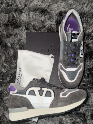 Valentino Garavani rockrunner skoe - Snygga sneakers från Valentino Garavani i grå mocka och mesh med vita och lila detaljer. Tjock sula med mönstrad undersida och stort V på sidan. Skön innersula och cool retrostil. Perfekta för dig som vill sticka ut med exklusiva detaljer. Allt fås med på bild. Skick 10/10 tveka ej på höra av er mvh