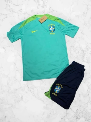 Brasilien fotbollsträningsset Nike - Snyggt träningsset från Nike med Brasilien-tema. Tröjan är turkos med gröna axelpartier, gul Nike-logga och Brasilien-emblem. Shortsen är mörkblå med Brasilien-logga och gröna detaljer. Perfekt för fotbollsträning eller match.