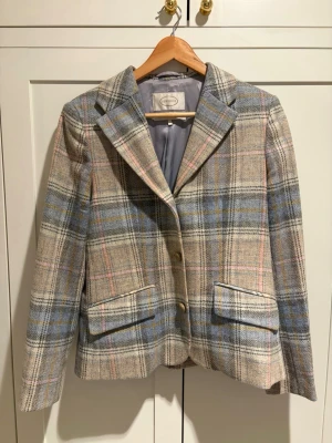 Rutig kavaj från Newhouse  - Snygg rutig kavaj från Newhouse  i beige, blått och rosa. Klassisk modell med två knappar framtill, ficklock och krage. Tillverkad i ullblandning med helfodrad insida. Perfekt för en stilren och trendig look. Knappt använd.