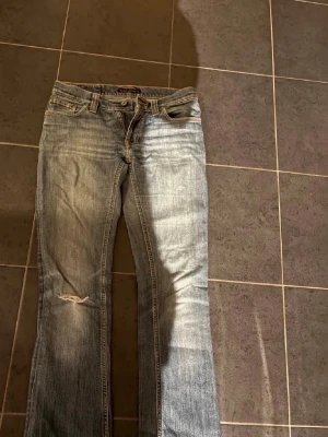 Blå bootcut jeans från Nudie Jeans - Säljer ett par ljusblå bootcut jeans från Nudie Jeans med slitningar och en liten reva på vänster ben. Klassisk femficksmodell med dragkedja och knapp. Snygga detaljer på bakfickorna och skön passform i mjukt denimtyg. Storlek 27/34, dock så är benen något avklippta så skulle säga att det är som en 32 nu.