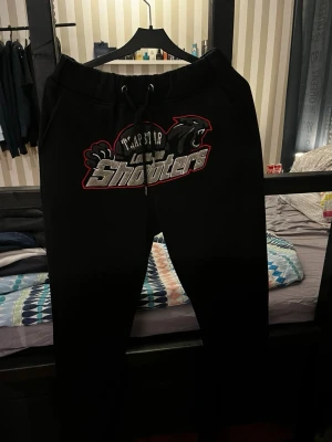 Trapstar mjukisbyxor - Svarta mjukisbyxor från Trapstar med stort broderat motiv och texten 'London Shooters' framtill. Byxorna har dragsko i midjan och är tillverkade i mjukt material som passar perfekt för en avslappnad stil.