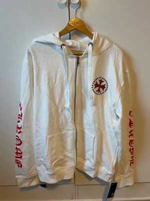 Vit Chrome Hearts zipper - Säljer en vit hoodie från Chrome Hearts med dragkedja, huva och stora röda tryck på både bröst, rygg och ärmar. Hoodien har kängurufickor och coola detaljer i form av logga och text. Perfekt för dig som gillar streetwear och vill sticka ut.