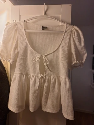 Vit blus med knyt från Gina Tricot - Supersöt vit blus från Gina Tricot med puffärmar och två knyt framtill. Blusen har en lätt rynkad struktur och volang nedtill som ger en romantisk vibe. Perfekt till jeans eller kjol för en trendig look.