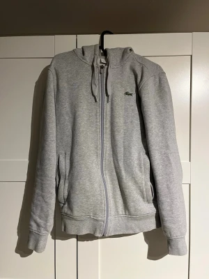 Grå zip-hoodie från Lacoste - Snygg grå hoodie från Lacoste med dragkedja framtill och klassisk krokodillogga på bröstet. Tröjan har huva med dragsko och två fickor på sidorna. Liten i storlek.