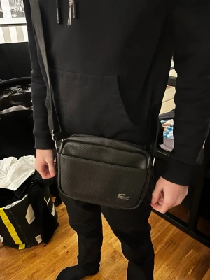 Lacoste väska - Snygg svart axelväska från Lacoste med justerbar rem och diskret krokodillogga på framsidan. Väskan har en rektangulär form och är tillverkad i tåligt syntetmaterial med struktur. Perfekt för att bära det viktigaste med stil.