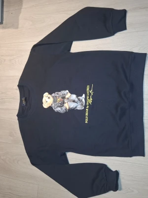 Svart sweatshirt Polo Bear Ralph Lauren - Cool svart sweatshirt från Ralph Lauren med den ikoniska Polo Bear-trycket på bröstet. Tröjan har rund halsringning och ribbade muddar vid ärmar och nederkant. Materialet känns mjukt och bekvämt, perfekt för en avslappnad stil.