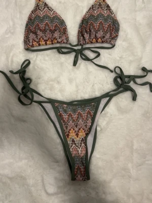 Mönstrad bikini med knyt i grönt - Snygg bikini med färgglatt mönster i grönt, rött, gult och rosa. Både överdel och underdel har knytband i mörkgrönt och trekantsformad topp. Perfekt för stranden eller poolen! Aldrig använd!