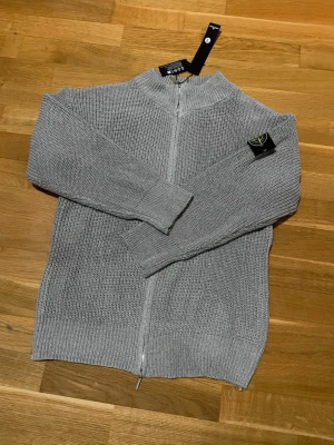 Grå stickad ziptröja Stone Island - Snygg grå stickad ziptröja från Stone Island med hög krage och ribbade muddar. Tröjan har dragkedja framtill och klassisk Stone Island logga. Säljer denna för den passa mig inte. Den är helt ny och taggar finns kvar. Komm med bud vid snabb affär.