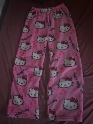 Hello Kitty Rosa pyjamasbyxor - Supermjuka och mysiga pyjamasbyxor i rosa fleece med Hello Kitty-motiv och små röda rosetter. Byxorna har resår i midjan och ett avslappnat, rakt ben. Perfekta för chillkvällar hemma eller när du vill vara extra bekväm.