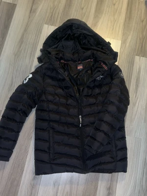 Svart dunjacka med huva från Napapijri - Snygg svart dunjacka från Napapijri med huva och dragkedja framtill. Jackan har quiltad design, två sidofickor och vita detaljer på ärmen. Perfekt för kalla vinterdagar och riktigt skön att bära.