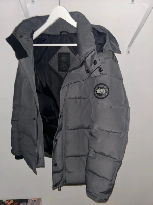 Canada Goose Wyndham - Säljer en grå dunjacka från Canada Goose  Jackan har en patch med logga på armen och stängs med dragkedja och knappar. Perfekt för kalla vinterdagar och riktigt skön att bära.