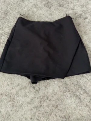 Svart skort med omlott-look - Stilren svart skort med omlott-design framtill och två dekorativa fickor bak. Klassisk och enkel modell som passar till många olika stilar. Materialet ser ut att vara ett mjukt tyg med lite struktur. Perfekt för dig som vill ha både kjol och shorts i ett plagg.