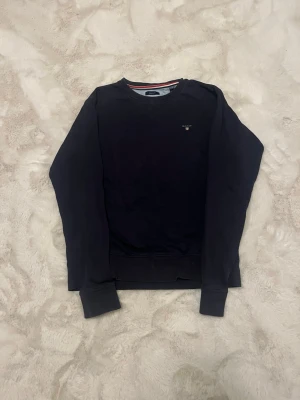 Mörkblå sweatshirt från Gant - Klassisk mörkblå sweatshirt från Gant med rund halsringning och diskret logga på bröstet. Tröjan har ribbade muddar vid ärmslut och nederkant samt en snygg detalj med röd och vit rand på insidan av kragen. Perfekt för en stilren och avslappnad look.