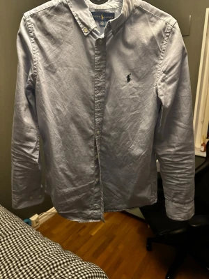 Ljusblå skjorta från Ralph Lauren - Klassisk ljusblå skjorta från Ralph Lauren med button-down krage och broderad logga på bröstet. Skjortan har långa ärmar och knäppning framtill. Perfekt för en stilren look.