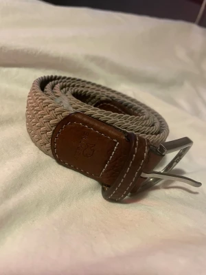 Beige flätat bälte från Selected - Snyggt beige flätat bälte från Selected med detaljer i brunt läder och silvrigt spänne. Perfekt accessoar för att lyfta din outfit med en stilren och modern touch. Bältet har klassisk design och passar till många olika looks.