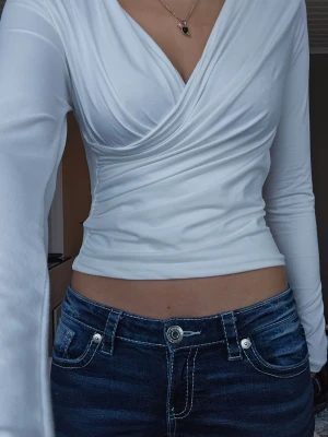 Vit omlottopp - Säljer en vit omlottopp med lång ärm och croppad passform. Toppen har en snygg v-ringning och sitter tight över kroppen. Perfekt att styla med jeans eller kjol för en trendig look.