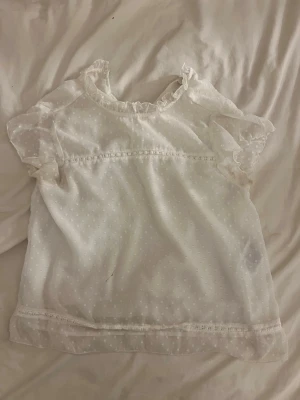 Vit prickig blus med volangärm - Söt vit blus i transparent material med små prickar och volangdetaljer vid ärmslut och hals. Blusen har rundad halsringning fram och v-ringning bak samt korta, luftiga ärmar. !!Om du köper välj gärna att hämta vid upphämtningsställe då det blir enklare för mig att posta på det viset!!