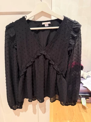 Svart blus med volanger och struktur - Snygg svart blus från Gina Tricot med volangdetaljer och prickig struktur i tyget. Blusen har v-ringning och långa, lätt transparenta ärmar med resår vid ärmslut. Perfekt för dig som gillar trendiga detaljer och en luftig känsla. Storleken är 164 och finns inte på plick så gissade bara.