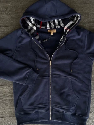 Marinblå hoodie från Burberry - Snygg marinblå hoodie med dragkedja från Burberry. Klassisk huva med det ikoniska rutmönstret på insidan, dragsko och två fickor framtill. Tillverkad i mjukt bomullsmaterial och perfekt för en avslappnad stil.
