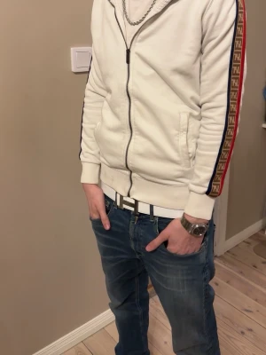 Vit Fendi hoodie med logga på ärmar - Snygg vit hoodie från Fendi med dragkedja och huva. Ärmarna har Fendi-logga i brunt, rött och blått längs sidorna. Hör av er för minsta fråga. Priset går att diskutera!