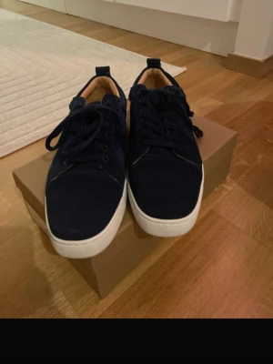 Mörkblå sneakers från Christian Louboutin - Snygga mörkblå sneakers från Christian Louboutin med klassisk röd sula och vit kant. Skorna är gjorda i mocka med snörning och har en rund tå. Insidan är beige och märkt med Louboutin Paris. Perfekt för dig som vill ha något extra till din stil.