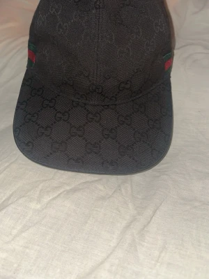 Svart keps från Gucci med monogram - Svart keps från Gucci med klassiskt GG-monogram över hela kepsen. På sidorna finns gröna och röda ränder som ger en lyxig känsla. Kepsen är i tygmaterial och har böjd skärm. Perfekt för dig som vill ha en stilren och trendig accessoar.