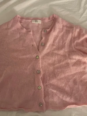 Ljusrosa stickad kofta med knappar - Söt ljusrosa stickad kofta med rund halsringning och sex vita knappar framtill. Koftan har en mjuk och tunn stickning, perfekt för lager-på-lager. Avslappnad passform och ribbade kanter vid hals och ärmslut. Får även med en extra knapp