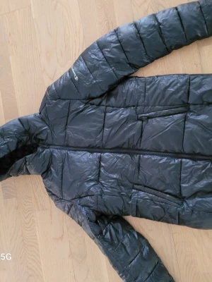 Dunjacka från Champion - Snygg mörkblå dunjacka från Champion med huva och dragkedja framtill. Jackan har två sidofickor med dragkedja och diskret Champion-logga på ärmen. Perfekt för kalla vinterdagar och ger en sportig look.