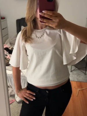 Vit croppad topp med vida ärmar - Trendig vit croppad topp med vida, korta ärmar och rund halsringning. Perfekt för dig som gillar en stilren och luftig look. Toppen har en enkel design utan synliga detaljer och passar till många olika outfits.