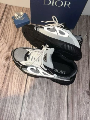 Dior grå och svarta sneakers - Snygga Dior sneakers i grått, svart och vitt med mesh och läderdetaljer. Skorna har en chunky sula och tydliga Dior-loggor på plösen och innersulan. Stilen är sportig och modern med vita snören och kontrasterande paneler.