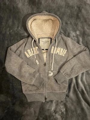 Grå hoodie från Abercrombie & Fitch - Mysig grå hoodie från Abercrombie & Fitch med dragkedja, broderad text på bröstet och vit teddyfoder i huvan. Tröjan har ribbade muddar, fickor framtill och tjocka snören i huvan. Perfekt för dig som gillar en avslappnad och trendig stil. Storlek M men skulle mer säga xs/s.     Pris kan diskuteras vid snabb affär!