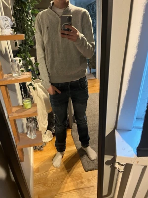 Ljusgrå quarter zip från Gant - Säljer en stilren ljusgrå tröja från Gant med quarter zip och liten broderad logga på bröstet. Tröjan har ribbade muddar och krage, perfekt för lager på lager. Tillverkad i mjukt bomullsmaterial och passar till det mesta.