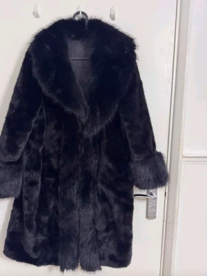 Svart fuskpäls kappa med bred krage - Snygg svart fuskpäls kappa med bred, fluffig krage och uppvikta ärmslut. Kappan har en rak passform och är helsvart med lyxig känsla. Perfekt för dig som vill ha en statement piece till vintern. Stängs framtill och har en klassisk siluett.
