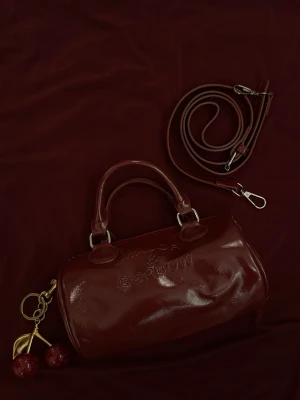 Mörkröd handväska - Cool mörkröd handväska i glansigt material från Major & Gordon. Väskan har rund form, dubbla handtag och avtagbar axelrem. Utsmyckad med ett körsbärsformad nyckelring i guld och rött. Perfekt för dig som gillar unika accessoarer.