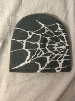 Grå mössa med vitt spindelnätsmönster - Snygg grå mössa med ett coolt vitt spindelnätsmönster runt hela. Mössan är slätstickad och har en klassisk, rundad form som sitter skönt på huvudet. Perfekt för dig som vill sticka ut lite extra under kalla dagar.
