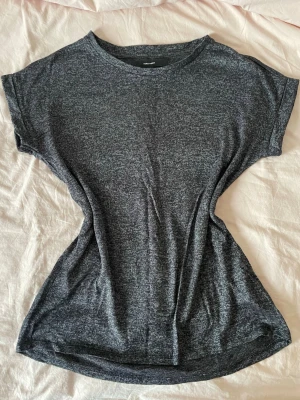 Mörkgrå t-shirt från Vero Moda - En basic mörkgrå t-shirt från Vero Moda i mjuk bomullsblandning. T-shirten har rund halsringning och korta ärmar, perfekt för en avslappnad och stilren look. Passformen är normal och tyget har en lätt melerad effekt. Den är lite genomskinlig dock som man kan se på tredje bilden.