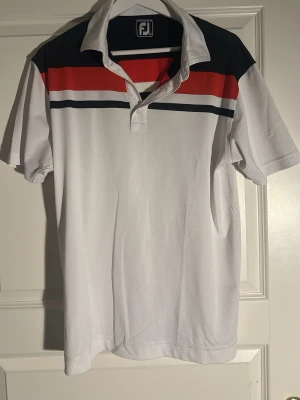 Vit FJ piké med röd och marinblå rand - Snygg vit pikétröja från FootJoy med breda ränder i rött och marinblått över bröstet och axlarna. Klassisk krage och knappslå framtill. Tillverkad i ett lätt och ventilerande material, perfekt för en sportig och avslappnad stil.