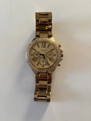 Guldfärgad klocka från Regal - Stilren guldfärgad klocka från Regal med länkarmband i metall och glittrande stenar runt urtavlan. Urtav the care har romerska siffror och flera små urtavlor för extra detaljerad look. Perfekt accessoar för att lyfta din outfit. Behöver nytt batteri