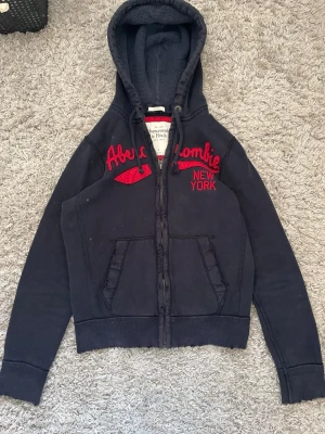 Mörkblå hoodie Abercrombie & Fitch - Mörkblå hoodie från Abercrombie & Fitch med dragkedja, huva och fickor framtill. Snygg röd brodyr med texten 'Abercrombie New York' på bröstet. Tillverkad i mjukt bomullsmaterial och har ribbade muddar vid ärmslut och nederkant. Perfekt för en avslappnad streetstyle.