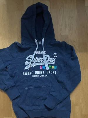 Mörkblå Superdry hoodie med tryck - Mörkblå hoodie från Superdry med vit logga och färgglada japanska tecken på bröstet. Klassisk känguruficka, ribbade muddar och justerbar huva med vita snören. Perfekt för en avslappnad streetstyle-look.