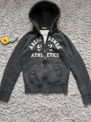Mörkgrå hoodie Abercrombie Athletics - Mörkgrå hoodie från Abercrombie & Fitch med dragkedja, huva med dragsko och stora fickor framtill. Tryck i vitt med texten 'Abercrombie Athletics 92' på bröstet. Skön collegekänsla och ribbade muddar vid ärmslut och nederkant.