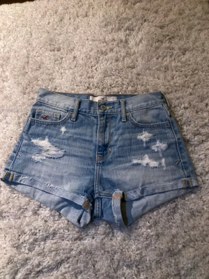 Ljusblå jeansshorts från Hollister - Säljer ett par ljusblå jeansshorts från Hollister med slitna detaljer och klassisk femficksdesign. Shortsen har normal passform, knappgylf och bälteshällor. Perfekta för sommaren och ger en avslappnad look.