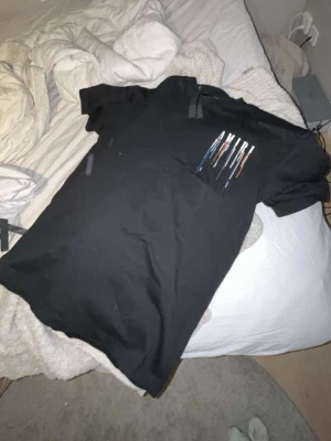 Svart AMIRI t-shirt med färgstänk - Säljer en svart t-shirt från AMIRI med stort tryck framtill där AMIRI-loggan syns tillsammans med färgstänk i blått, orange och vitt. T-shirten har korta ärmar och en klassisk passform. Perfekt för dig som vill ha en statement tee med cool design.