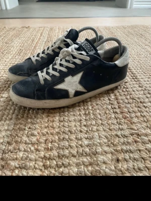 Svarta Golden Goose sneakers med stjärna - Snygga svarta sneakers från Golden Goose med vit stjärndetalj på sidan och vita skosnören. Skorna har en låg silhuett och vit sula. Tillverkade i läder med klassisk GGDB-logga på plösen och sidan. Perfekta för en trendig och avslappnad stil.