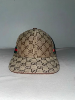 Gucci keps med GG-mönster - Snygg keps från Gucci i beige och brunt med klassiskt GG-monogram över hela kepsen. Kepsen har böjd skärm och detaljer i grönt och rött på sidorna. Perfekt accessoar för att lyfta din streetwear-look.